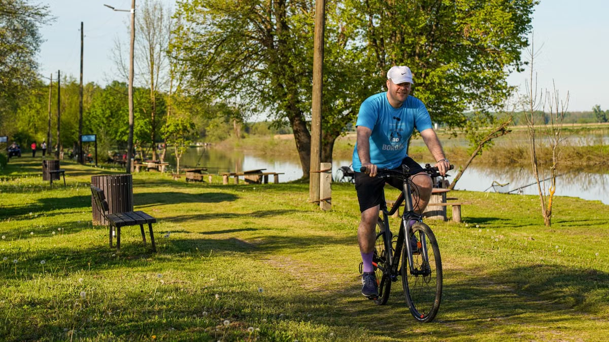 Adult man cycling along a lakeside path in a sunny park setting, surrounded by greenery and benches.