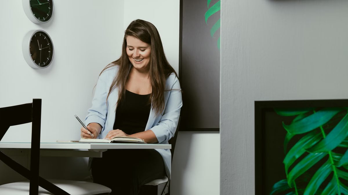 Cheerful young female journalist in casual clothes smiling while taking notes in planner during work in creative office